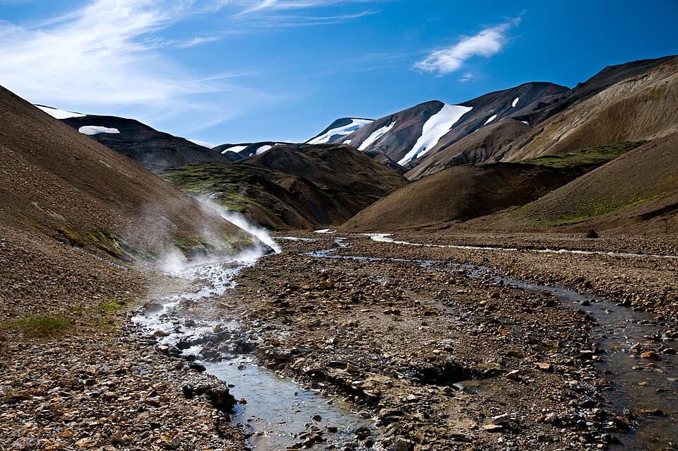 A highland valley in Iceland in summer with snow patches on the slopes, a stream running through gravel, and steam rising from geothermal ground
