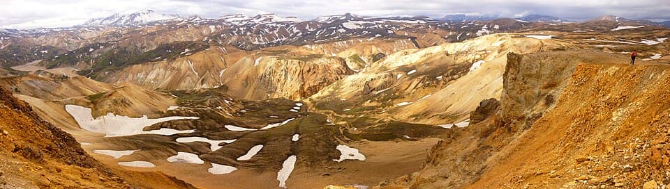A wide panorama of the Icelandic highlands with snow patches and braided river valleys, with a single hiker visible on the ridge at the right for scale