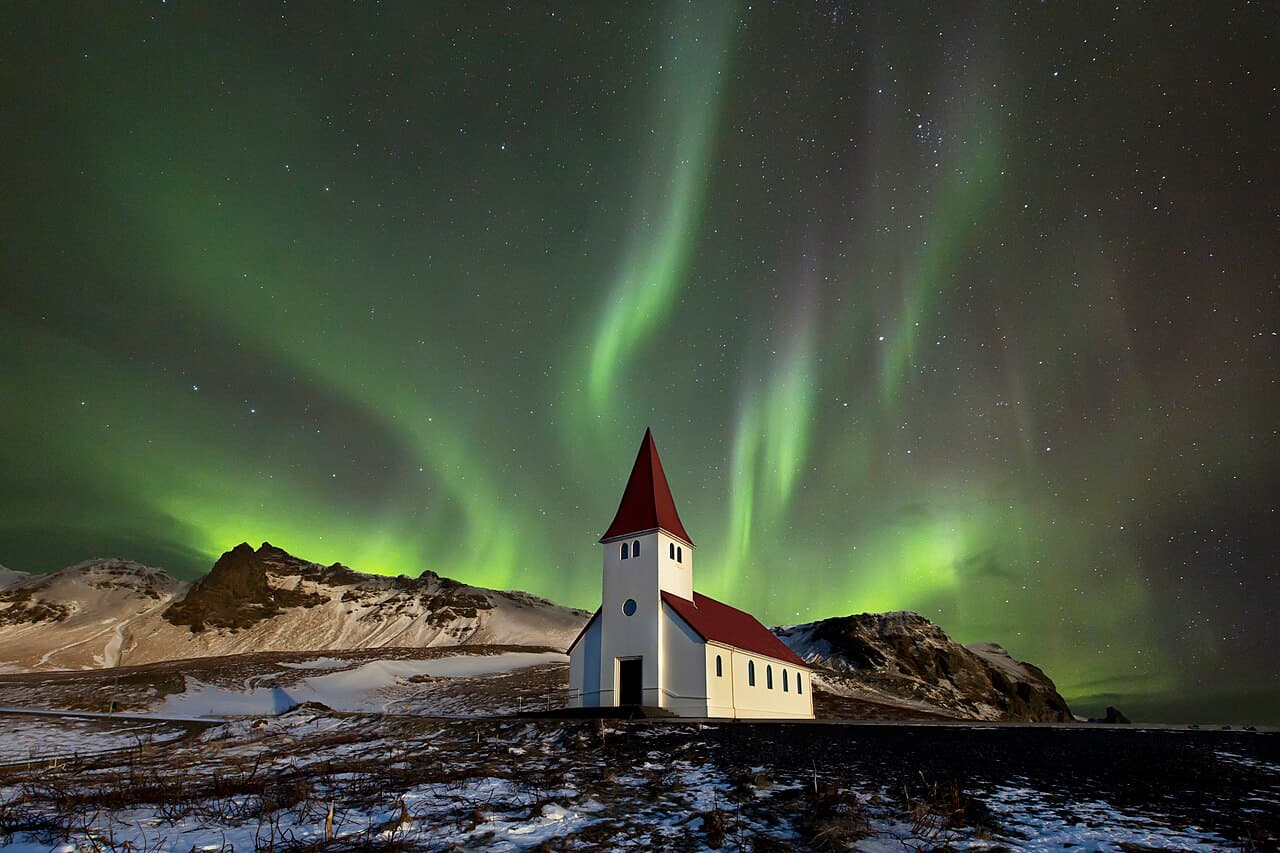 Green aurora bands over the church at Vík with snow-covered mountains behind