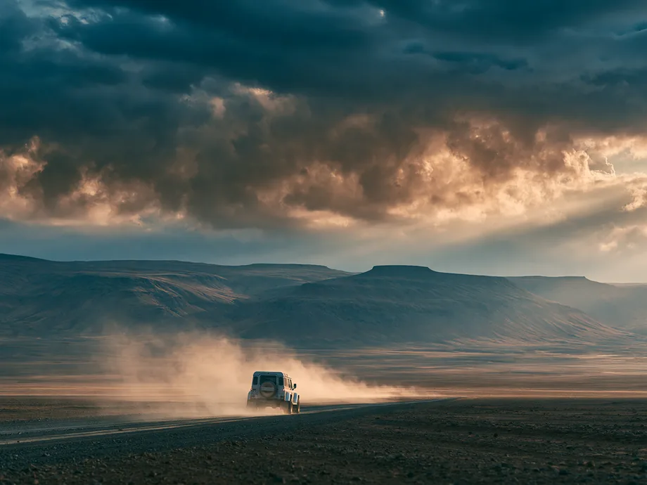 A 4x4 trailing dust on a highland gravel road under storm light