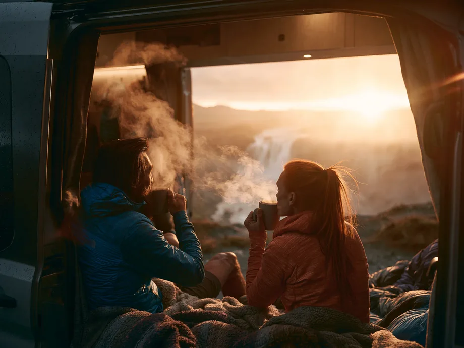 A couple with coffee in a camper van doorway facing a sunrise waterfall