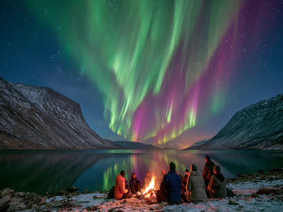 A small group around a campfire on a fjord shore under the northern lights